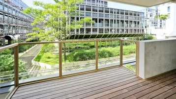 Geräumiger Balkon mit Holzboden und Glasgeländer, bietet Blick auf den begrünten Innenhof und moderne Gebäude.