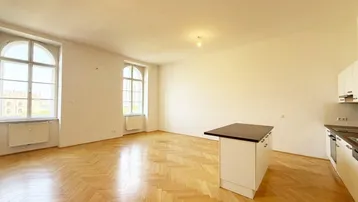 Helles Zimmer mit Fischgrätparkett, hohen Fenstern und integrierter Küchenzeile mit Insel.