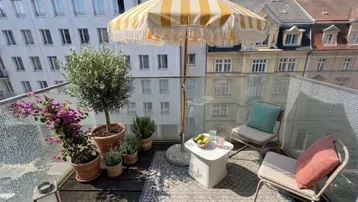 Möblierter Balkon mit Sonnenschirm, Pflanzen und Sitzgelegenheiten, umgeben von Stadtgebäuden.