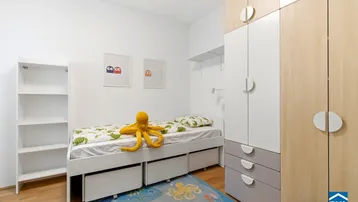 Helles Kinderzimmer mit Einzelbett, Stauraum und einem bunten Teppich.