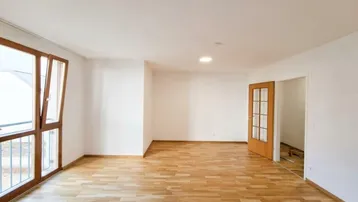 Helles, leeres Wohnzimmer mit Holzboden, großen Fenstern und Zugang zu einem Balkon.