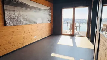 Heller Raum mit Holzwänden, großem Fenster und Zugang zu einem Außenbereich mit Seeblick.