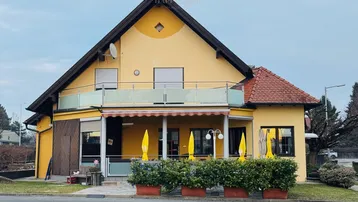 Gelbes Gebäude mit rotem Ziegeldach, Balkon und überdachtem Außenbereich mit Sonnenschirmen.