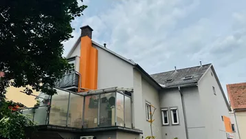 Gebäudeansicht mit Balkon und verglastem Anbau, umgeben von Bäumen und Hecken.