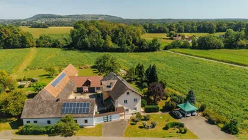 Weitläufiges Anwesen mit Photovoltaikanlage, umgeben von grünen Feldern und Wäldern unter strahlendem Himmel.