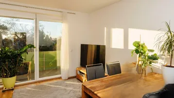 Heller Wohn- und Essbereich mit Holzboden, großem Fenster und Zugang zum Balkon.