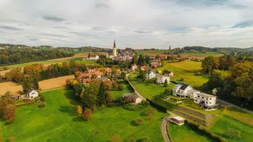 Luftaufnahme eines malerischen Dorfes mit Kirche, umgeben von grünen Feldern und herbstlichen Bäumen.