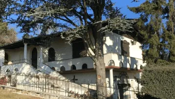 Traditionelles Haus mit weißer Fassade, dunklen Fensterläden und umgebenden Bäumen unter blauem Himmel.