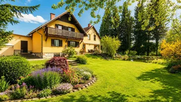 Gelbes Haus mit Holzbalkon, umgeben von einem gepflegten Garten mit Blumenbeeten, Rasenfläche und einem Teich.