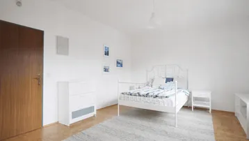 Helles Schlafzimmer mit weißem Metallbett, Holzparkettboden und einem großen Teppich.