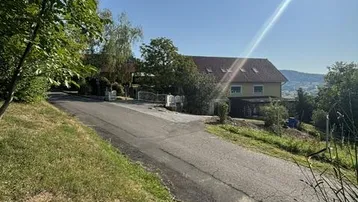 Blick auf ein Wohnhaus am Hang mit sonniger Umgebung und einer Zufahrtsstraße.