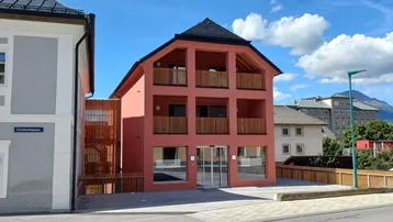 Ansicht eines modernen, rosafarbenen Gebäudes mit mehreren Balkonen und großen Fenstern in einer städtischen Umgebung.