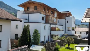 Modernes Mehrfamilienhaus mit weißer Fassade, Holzverkleidung, Balkonen und einem gepflegten Garten mit Bergblick.
