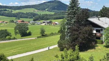 Haus mit Balkon in ländlicher Umgebung, umgeben von grünen Hügeln und Wäldern unter blauem Himmel.