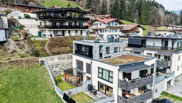 Panoramablick auf moderne Wohngebäude in einer grünen Berglandschaft mit bewaldeten Hängen.