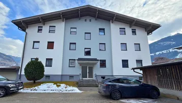Modernes Mehrfamilienhaus mit heller Fassade und Satteldach, umgeben von winterlicher Berglandschaft.