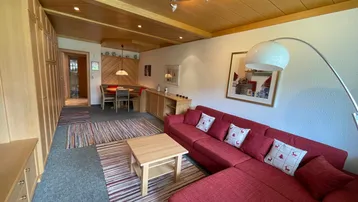 Geräumiger Wohnbereich mit rotem Ecksofa, Teppichboden und Essbereich im Hintergrund.