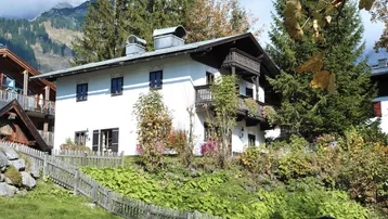 Traditionelles Alpenhaus mit Balkonen und Garten auf einem Hang, umgeben von Bäumen und Bergen.
