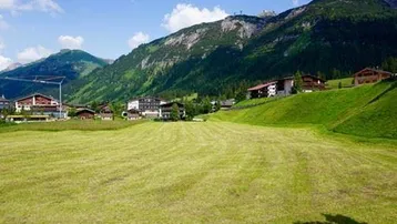 Grüne Wiesen und alpine Landschaft mit Häusern und majestätischen Bergen im Hintergrund an einem sonnigen Tag.