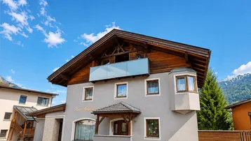 Traditionelles Haus mit Holzbalkonen und grauer Fassade unter blauem Himmel mit Bergblick.