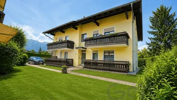 Gelbes Mehrfamilienhaus mit traditionellen Holzbalkonen, umgeben von einem gepflegten Rasen und Bergblick.