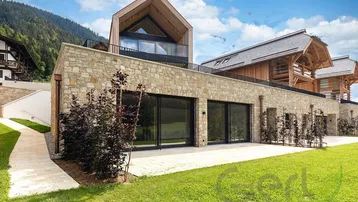 Moderne Architektur mit Natursteinfassade und Holzverkleidung, umgeben von grüner Landschaft und Bergen.