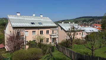 Mehrere Wohnhäuser mit Gärten und Spielplatz in ländlicher Umgebung unter blauem Himmel.