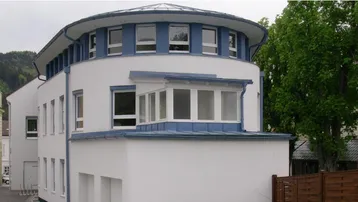 Modernes Gebäude mit runder Architektur, weißer Fassade und blauen Akzenten unter bewölktem Himmel.