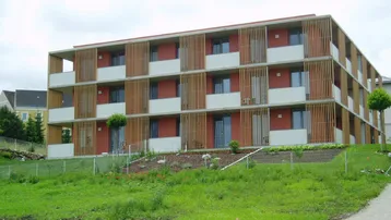 Modernes Mehrfamilienhaus mit roter Fassade und vertikalen Holzlamellen, umgeben von Grünflächen.