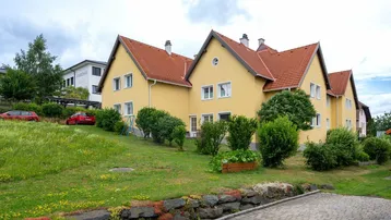 Gelbes Mehrfamilienhaus mit rotem Ziegeldach und gepflegtem Vorgarten auf einem Hügel.