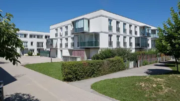Modernes Wohngebäude mit weißer Fassade, großen Fenstern und begrünten Außenbereichen unter blauem Himmel.