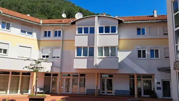 Ansicht des Gebäudekomplexes mit Innenhof und gepflastertem Bereich unter blauem Himmel, umgeben von grünen Hügeln.