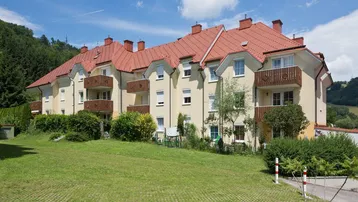 Mehrfamilienhaus mit mehreren Balkonen und gepflegter Grünfläche in hügeliger Umgebung.