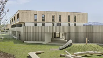 Gemeinschaftlicher Gartenbereich mit Spielplatz, Rutsche und Holzelementen, umgeben von Grünflächen und Bergblick.