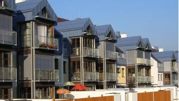 Reihen moderner Stadthäuser mit blauen Dächern, Balkonen und Rollläden unter klarem Himmel.