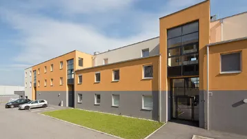 Moderne Wohnanlage mit orange-grauer Fassade und Parkplätzen vor dem Gebäude unter blauem Himmel.