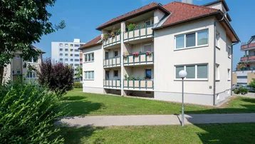 Mehrfamilienhaus mit hellbeiger Fassade, roten Ziegeldächern und Balkonen in einer grünen Umgebung.