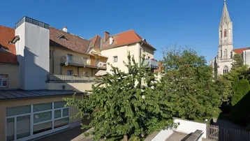 Ansicht des Gebäudes mit umliegender Grünfläche und Kirchturm im Hintergrund.