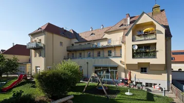 Mehrfamilienhaus mit großem Gemeinschaftsgarten, Spielplatz und Balkonen unter blauem Himmel.