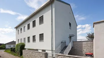 Ansicht eines mehrstöckigen Gebäudes mit heller Fassade, vielen Fenstern und einer Außentreppe.