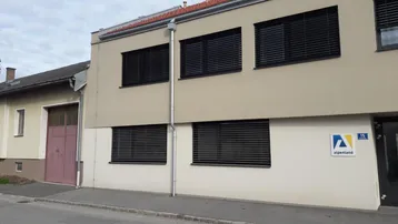 Modernes Gebäude mit beiger Fassade, schwarzen Jalousien und einem Firmenlogo an der Wand.