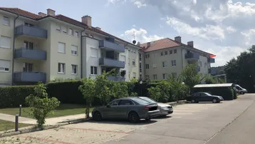 Mehrfamilienhaus mit Balkonen und Außenparkplätzen unter bewölktem Himmel, umgeben von Grünflächen.