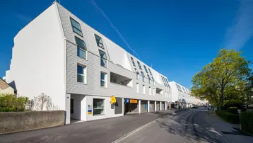 Modernes Gebäude mit weißer Fassade, Schrägdachfenstern und Geschäften im Erdgeschoss an einer Straße.
