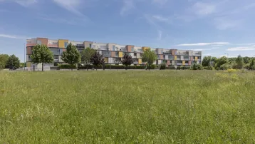Modernes Mehrfamilienhaus mit farbenfroher Fassade, umgeben von einer weiten Grünfläche unter blauem Himmel.