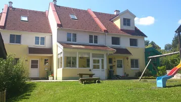 Reihenhäuser mit gemeinsamem Gartenbereich und einem kleinen Spielplatz unter blauem Himmel.