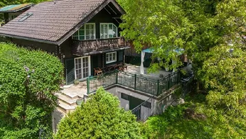 Traditionelles Haus mit großer Terrasse, umgeben von üppigem Grün und Bäumen.