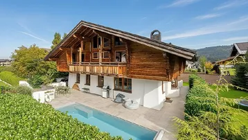 Traditionelles Chalet mit Außenpool, Bergblick und gepflegtem Garten unter blauem Himmel.