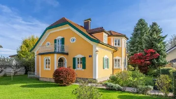 Gelbes Einfamilienhaus mit grünem Dach und gepflegtem Vorgarten unter blauem Himmel.