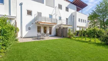 Hinteransicht des Hauses mit großer Rasenfläche, möblierter Terrasse und Balkonen.