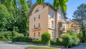 Historisches Gebäude mit mehreren Balkonen und gepflegter Hecke in grüner Umgebung.